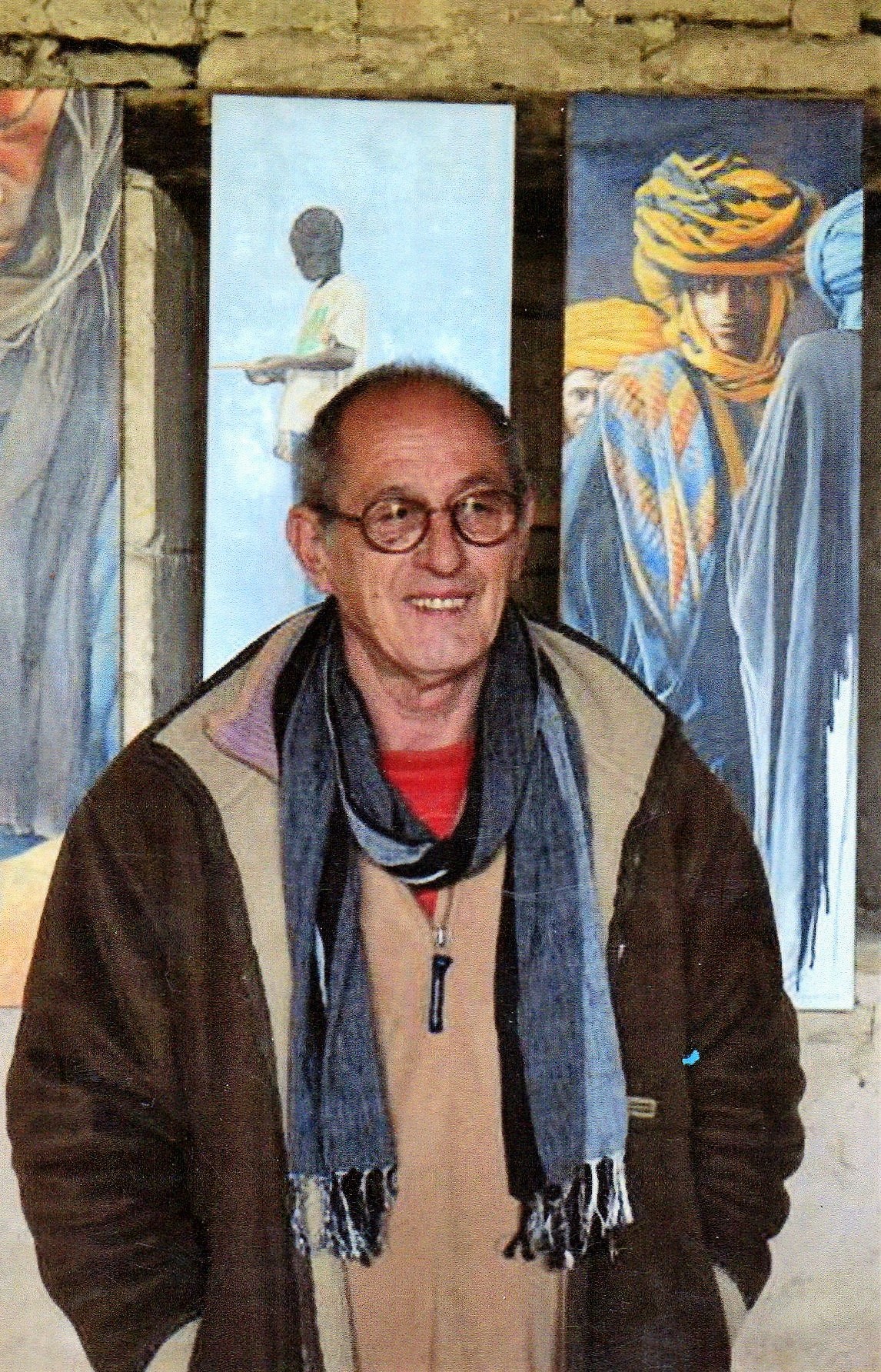 Jean-Louis Aparicio devant trois de ses grandes toiles verticales, des figures enturbannées et un enfant, accrochées contre un mur de pierre.